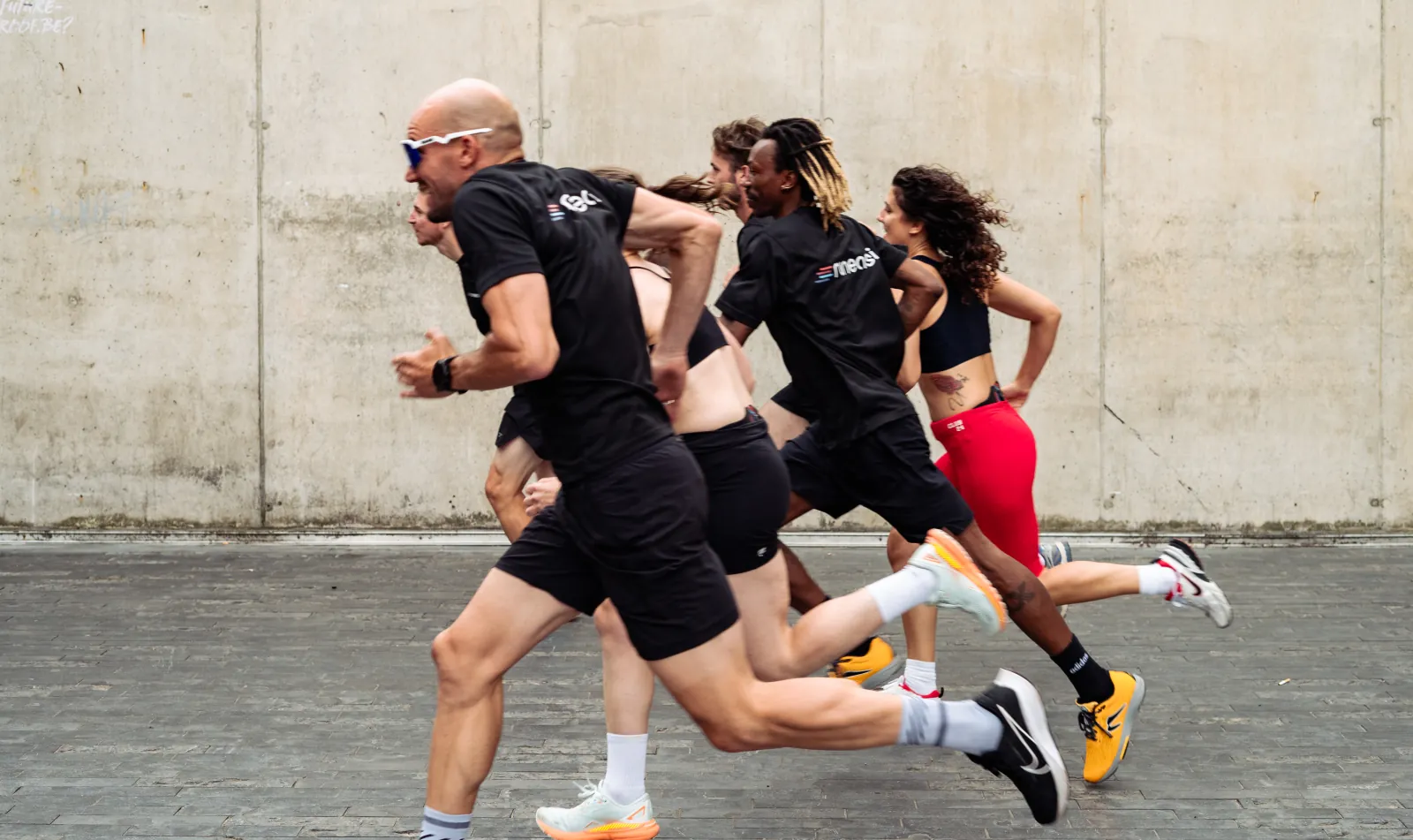 A group of runners in Runeasi shirts sprinting past a concrete wall