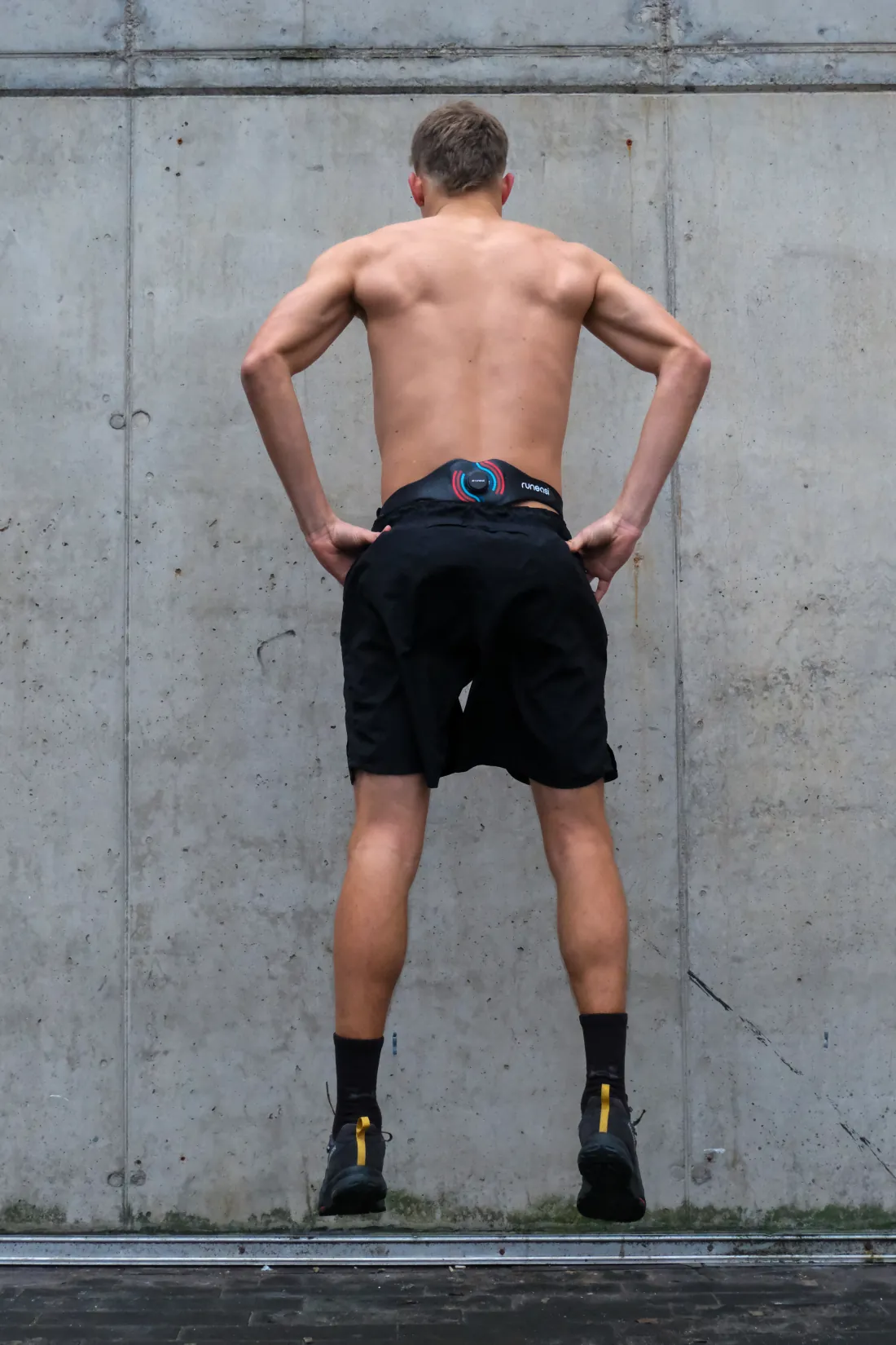 A runner stands facing a concrete wall with hands on hips, wearing the Runeasi belt