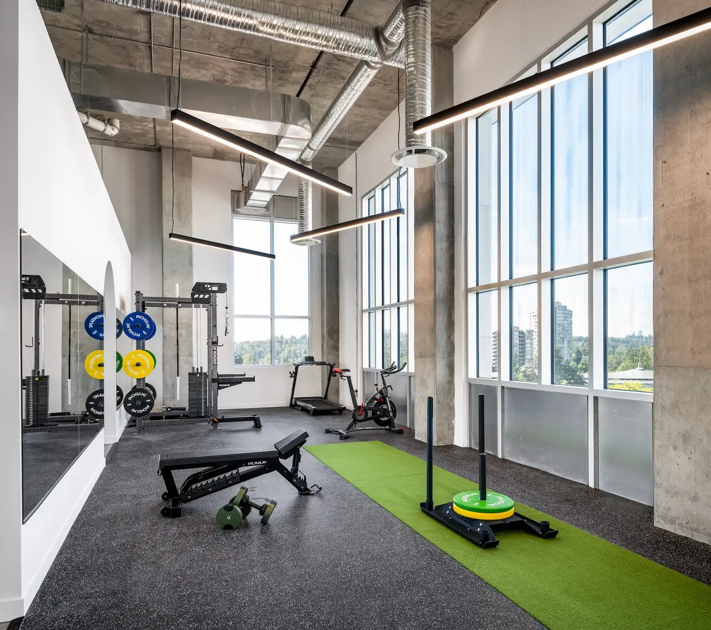 Inside Launch Rehab Lougheed strength floor — Rogue power squat rack stacked with blue, yellow, and green bumper plates against the mirror wall on the left with the arched mirror reveal beside it, Rogue adjustable bench centred in the foreground with green dumbbells on the rubber floor, Inspire treadmill and stationary bike along the floor-to-ceiling windows in back with Lougheed corridor views of distant towers and trees, Rogue weighted sled on the green turf strip on the right, exposed silver HVAC ducts and black linear LED pendants overhead