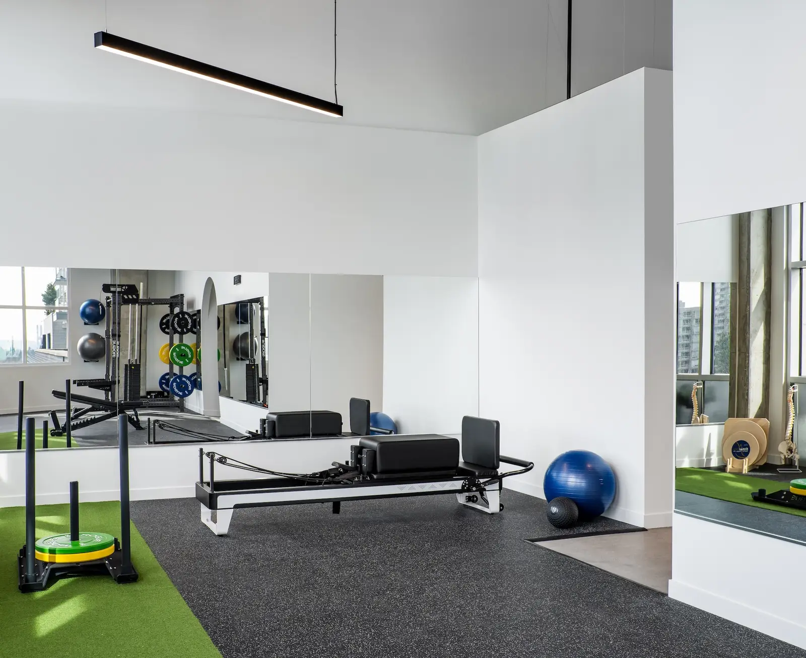 Inside Launch Rehab Lougheed Pilates and gym floor — Balanced Body reformer centred on the dark rubber gym floor with green turf strips on either side, mirror wall behind reflecting the Rogue squat rack stacked with colourful bumper plates, blue stability ball and 10lb slam ball against the white wall, black linear LED pendant overhead, and the arched mirror reveal showing the strength-training area beyond