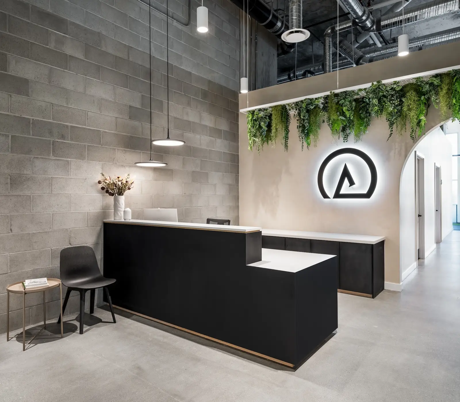 Inside Launch Rehab Lougheed — black reception desk with white quartz countertop and warm-wood reveal trim, side chair and metal side table on the left in front of a grey cinder-block wall, twin black pendant lights overhead, and the backlit Λ logo on a warm-beige plaster wall under the hanging plant wall with the arched corridor opening to the treatment hallway
