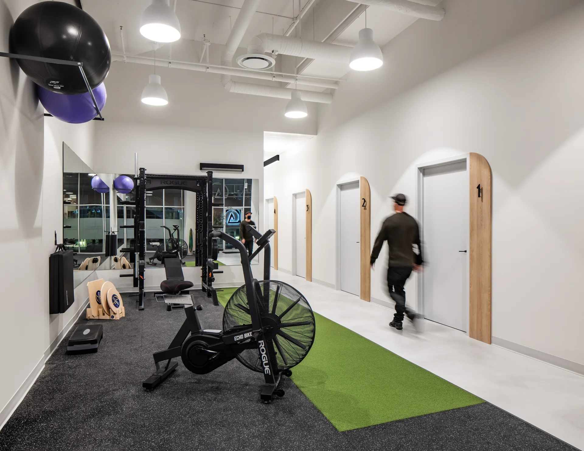 Gym floor at Launch Rehab New Westminster with Rogue Echo bike, squat rack, mirrors, and astroturf strip leading past numbered treatment rooms