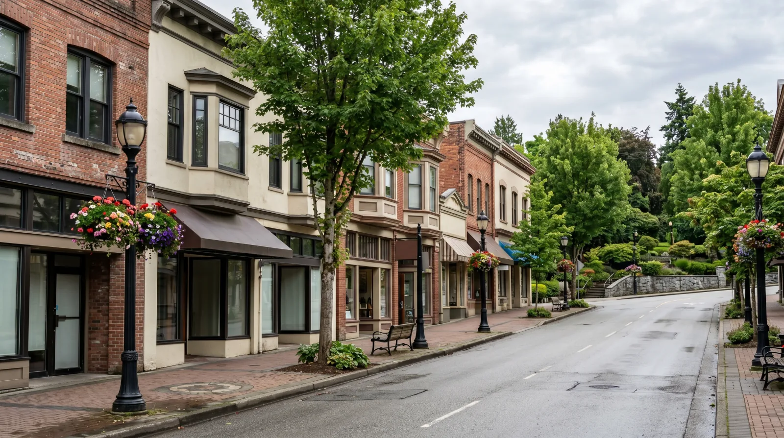 An established hilltop commercial street of older brick storefronts with flower baskets and a leafy park beyond — evoking Uptown New Westminster around Sixth Street.