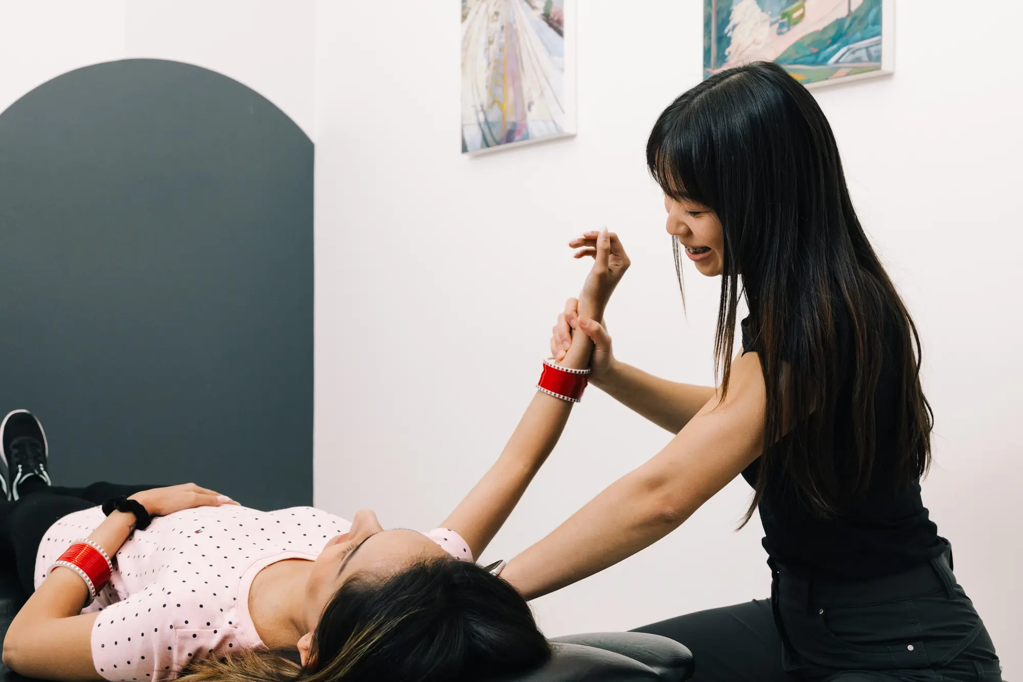A Launch Rehab physiotherapist working through a shoulder mobilisation with a patient on the treatment table — both wearing matching red beaded bracelets — under colourful framed coastal paintings, beside the studio's signature dark slate arched accent wall