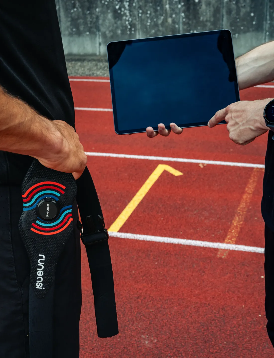 A Runeasi belt hangs from a hand on a running track while another person holds a tablet