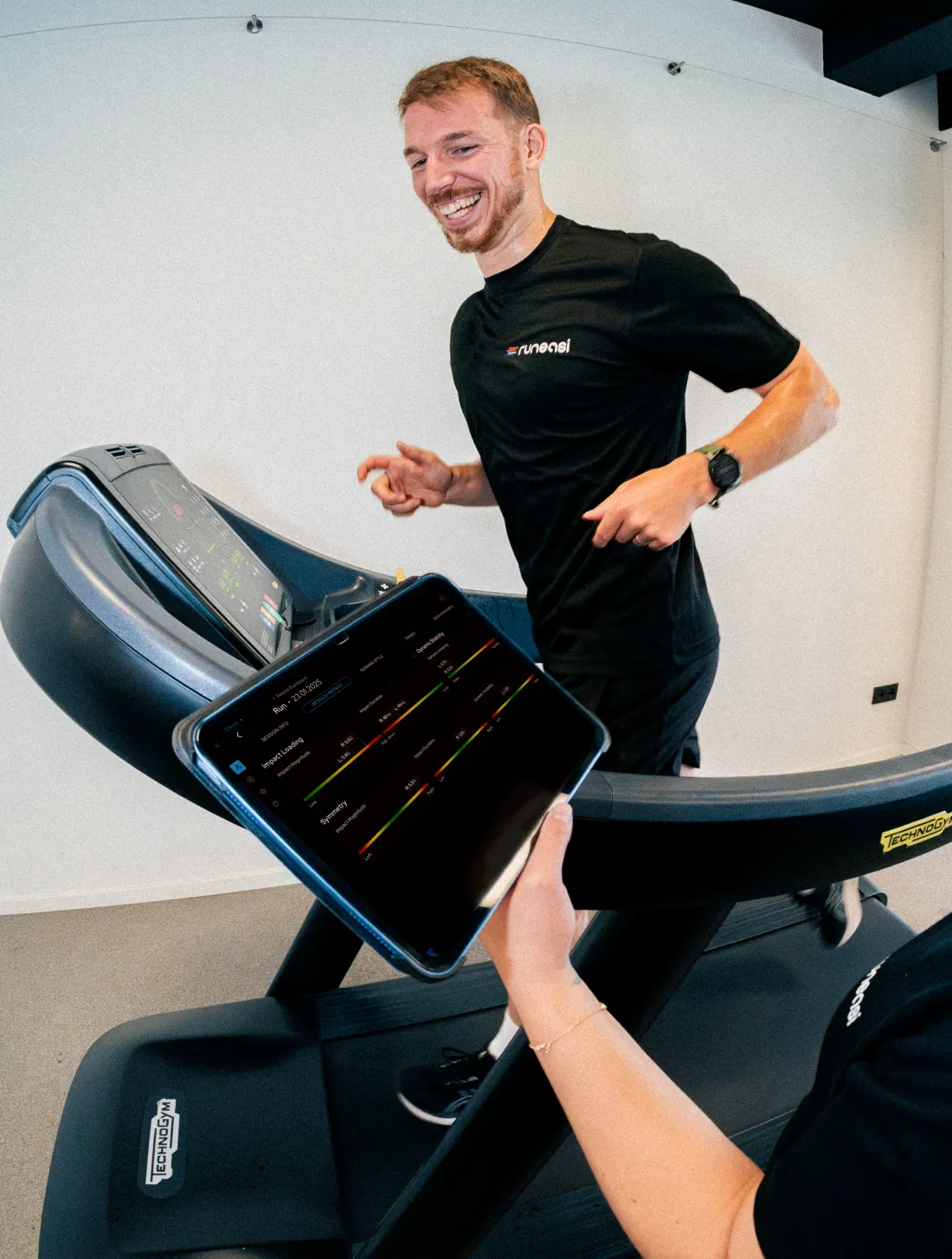 A runner laughs on a treadmill while a tester holds up a tablet with live data
