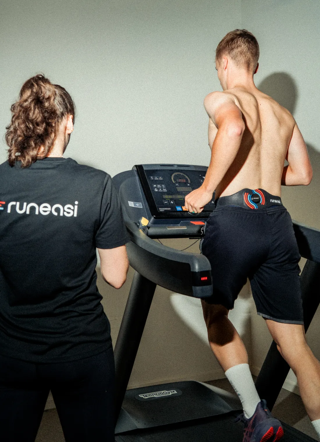 A tester in a Runeasi shirt watches a runner on a treadmill who wears the belt